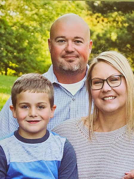 A smiling family of three—a man, a woman in glasses, and a young boy—pose together outdoors in front of green trees and grass.