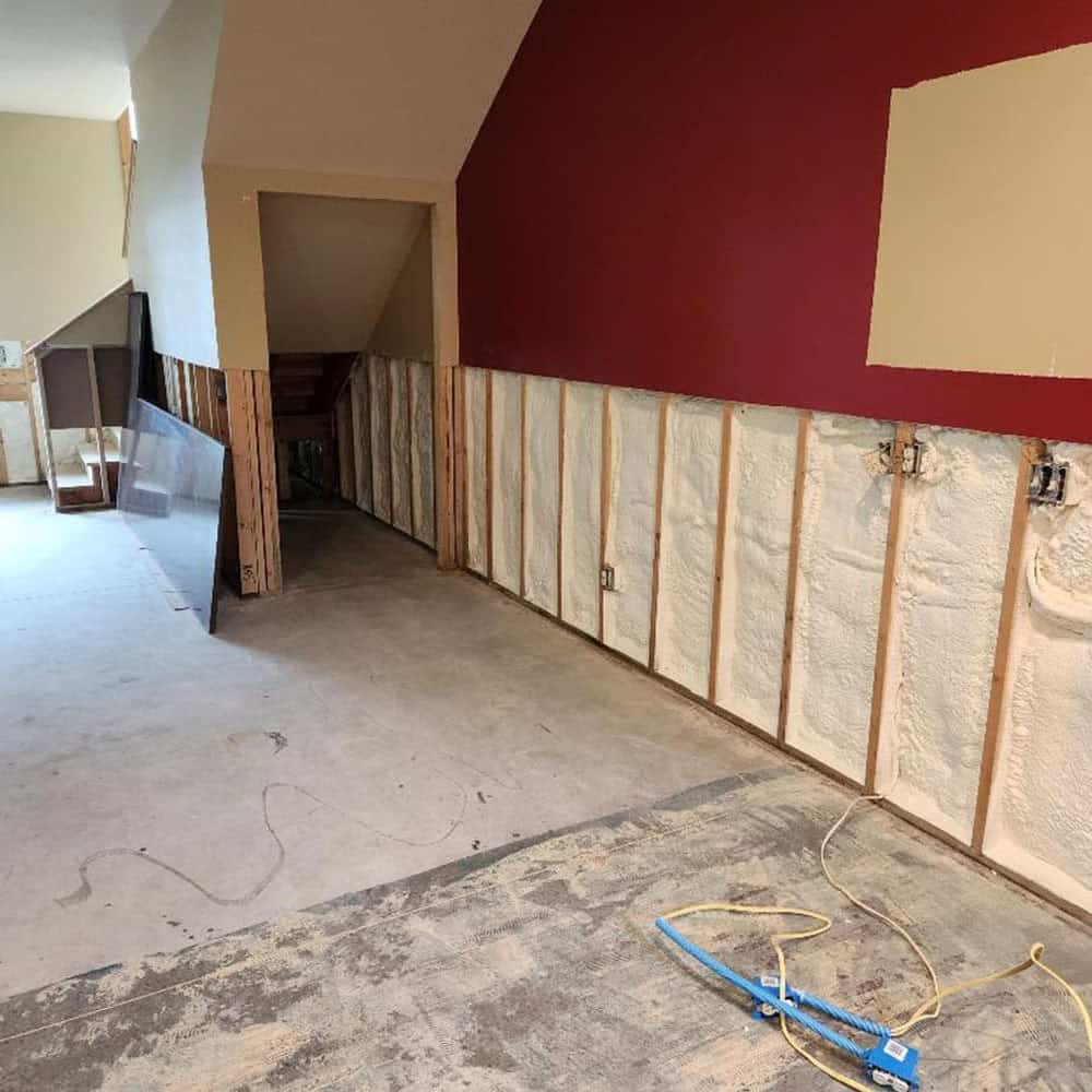 A room under renovation with exposed wall studs and fresh spray foam insulation. The floor is bare, wires are strewn about, and construction materials are scattered throughout the space.