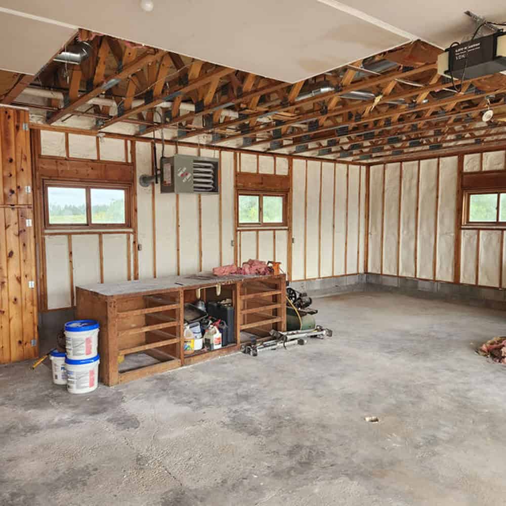 A spacious garage with exposed ceiling beams, spray foam insulated walls, concrete floor, a wooden workbench, paint cans, and tools. Two small windows let in natural light.