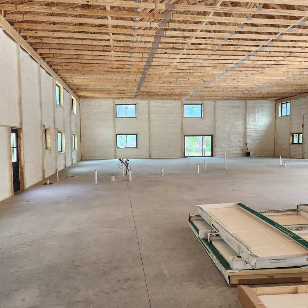 Large unfinished building interior with exposed wooden ceiling beams, spray foam insulated walls, multiple windows, concrete floor, and some construction materials and pipes scattered throughout the space.