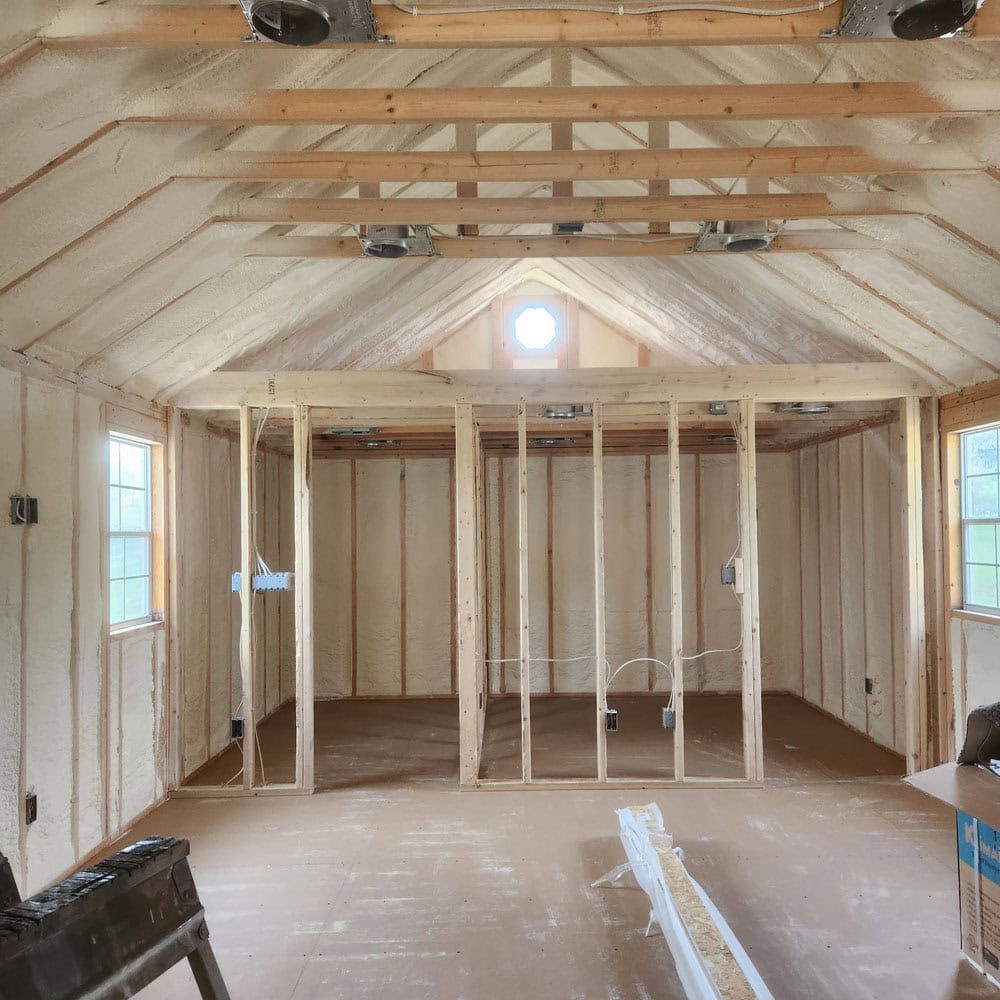 Interior of a house under construction with exposed wooden framing and spray foam insulation on the walls and ceiling. Windows let in natural light; electrical wiring is visible, and building materials are scattered on the floor.