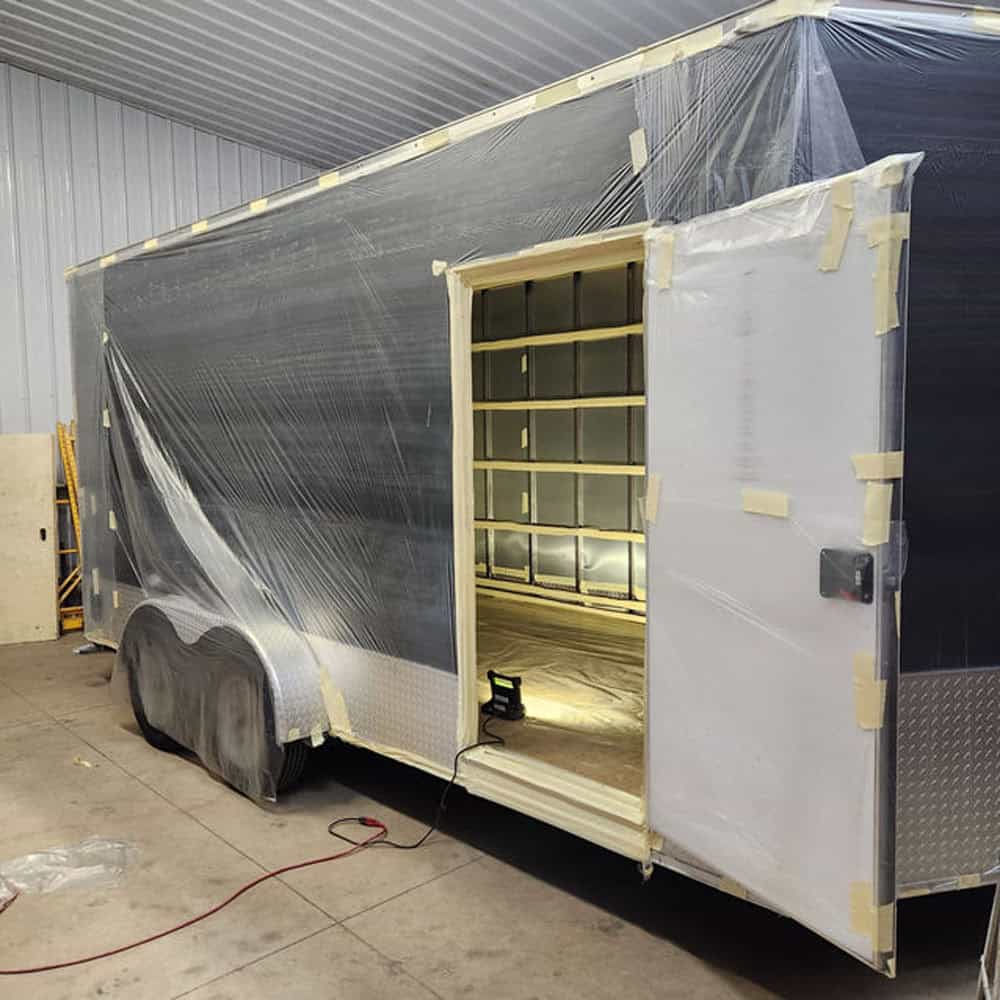 A large enclosed trailer is partially covered in clear plastic sheets and masking tape, parked inside a garage. The side door is open, revealing the unfinished interior. A work light is placed on the floor near the entrance.