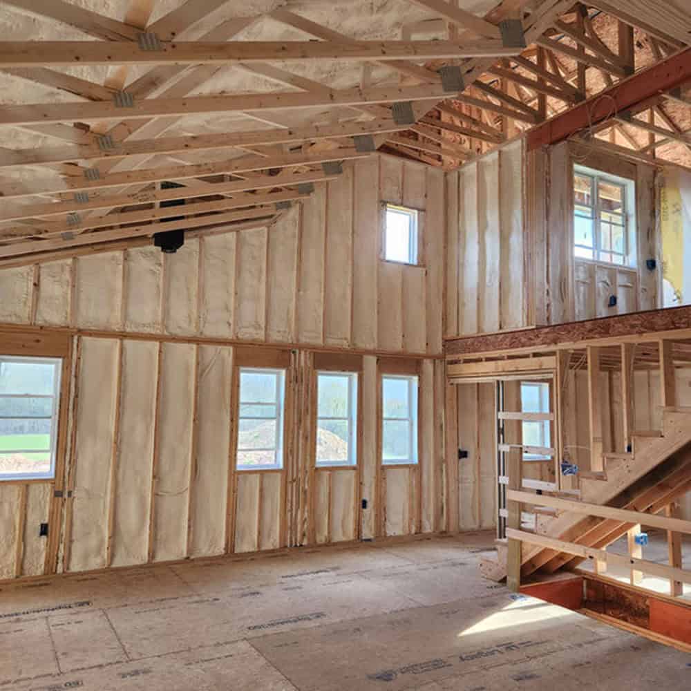 Interior of a house under construction, showing exposed wooden framing, foam insulation on walls, several windows, and a staircase leading to an unfinished second floor. Natural light enters through the windows.