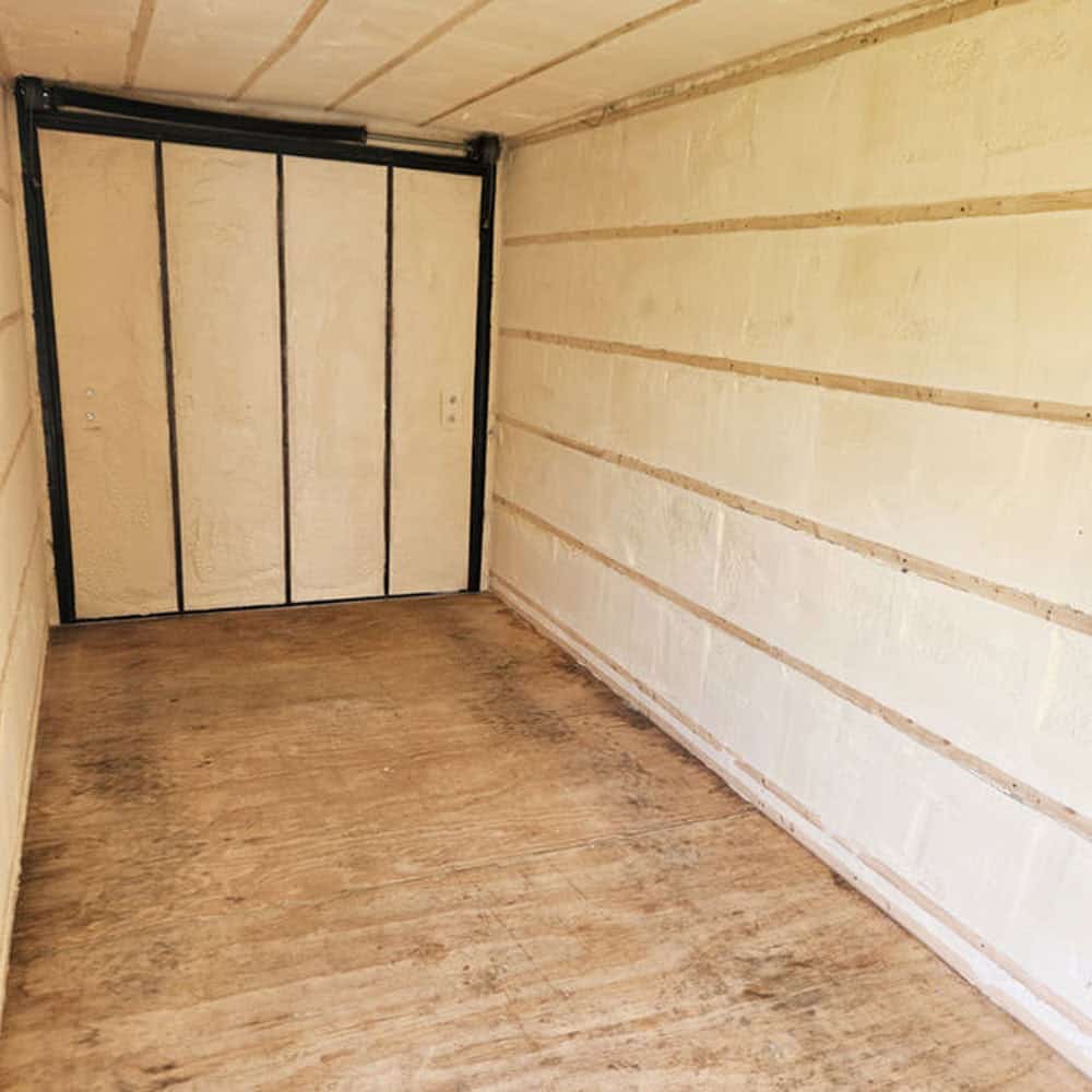 An empty, narrow room with wooden flooring and off-white insulated foam walls and ceiling. At the end, there are double doors with a metal frame. The space appears unfinished and utilitarian.