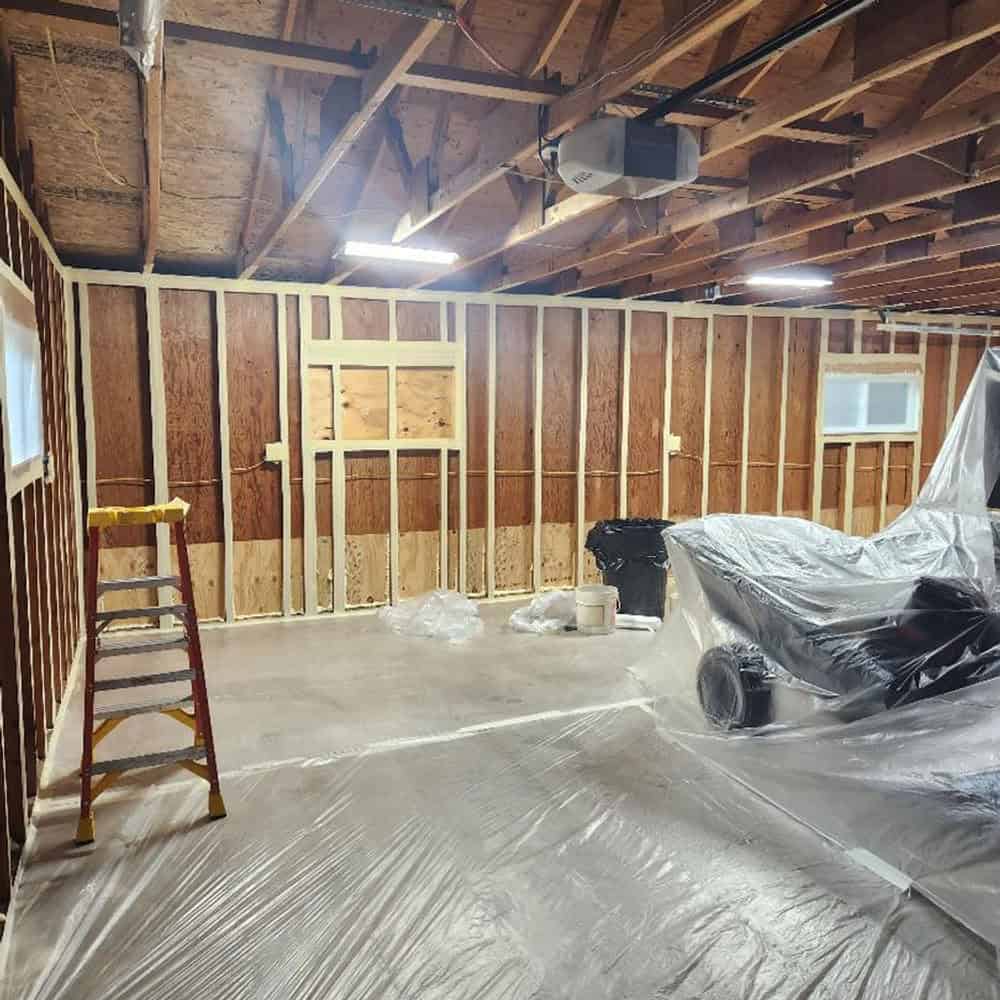 A garage under renovation with exposed wall framing, a ladder, and plastic sheets covering the floor and a parked car. Construction materials and tools are visible in the space.