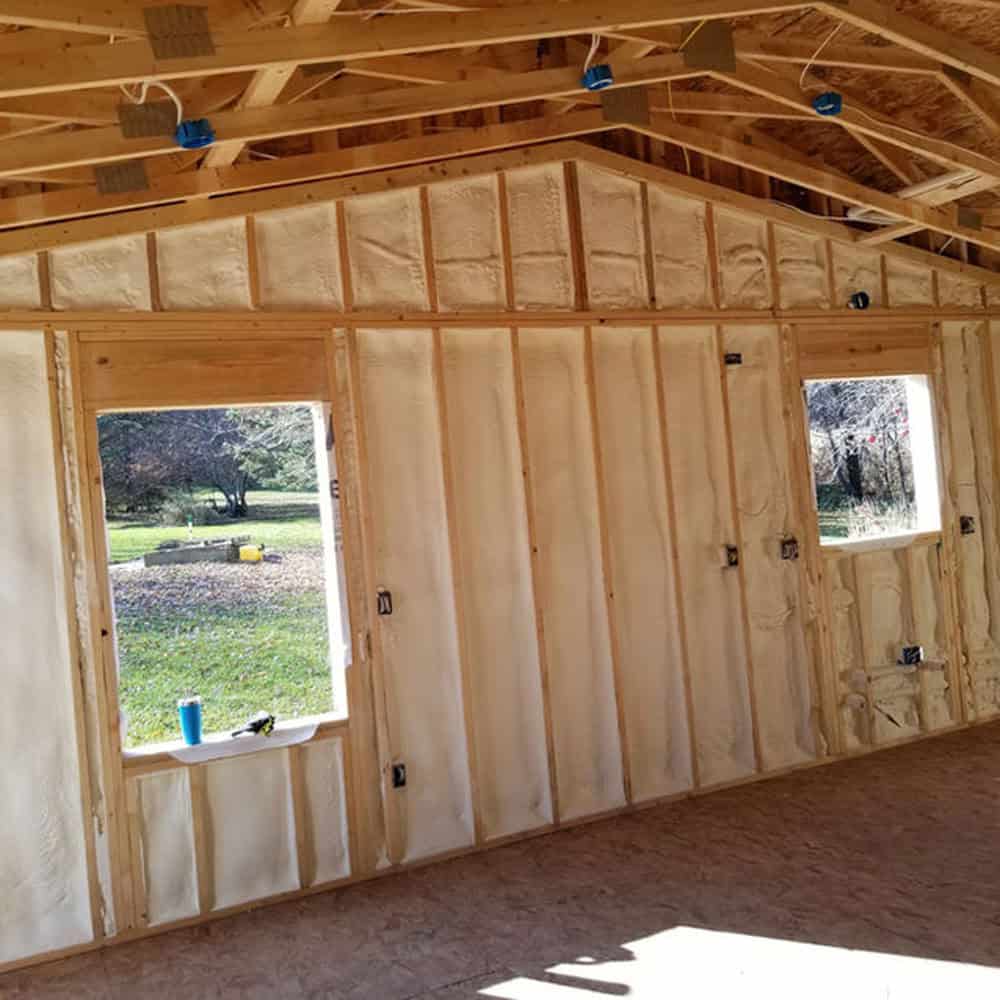 Interior view of a wooden structure with exposed framing and spray foam insulation on the walls, two large rectangular windows, and a plywood floor. Green grass and trees are visible outside through the windows.