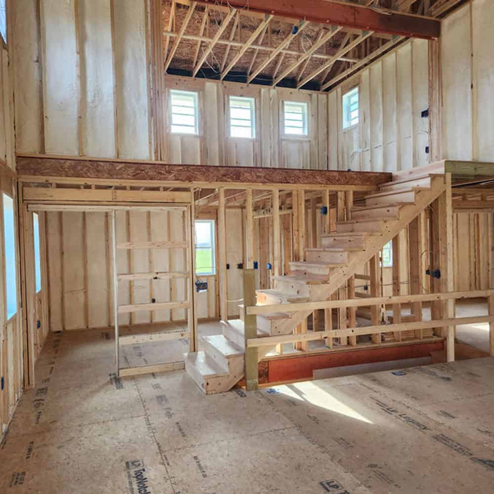 Interior of a house under construction, showing exposed wooden framing, an unfinished staircase leading to a loft area, and multiple small windows along the upper wall. No drywall or finishes are installed.