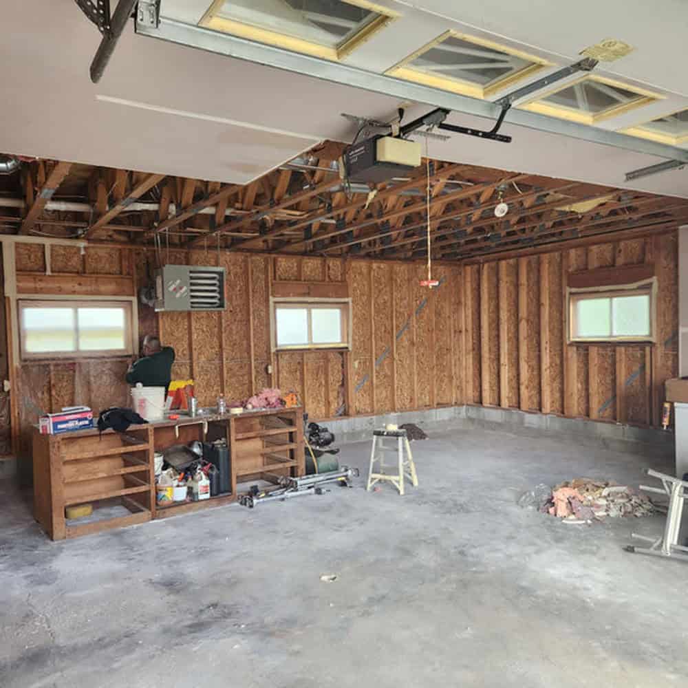 A garage with exposed wooden studs and ceiling, a workbench with tools and supplies, a step ladder, and building materials scattered on the concrete floor. A person is working near the wall.