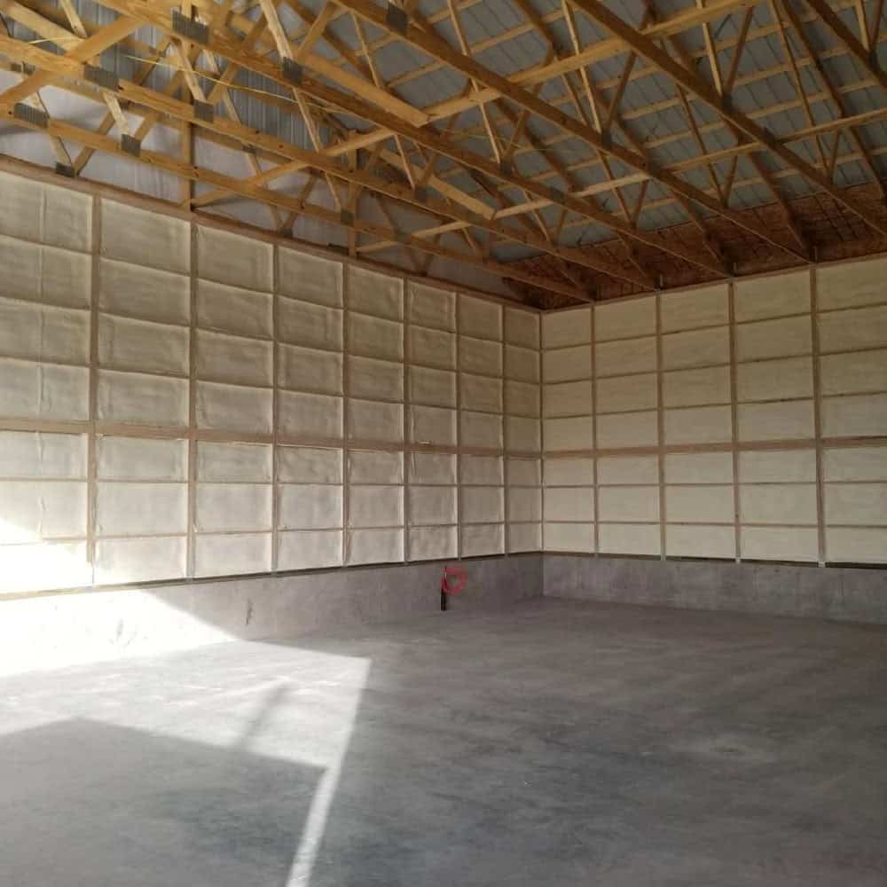 An empty warehouse with wooden roof trusses, concrete floor, and walls lined with white insulation panels. A single red fire extinguisher is mounted on the far wall. Sunlight enters from the left.