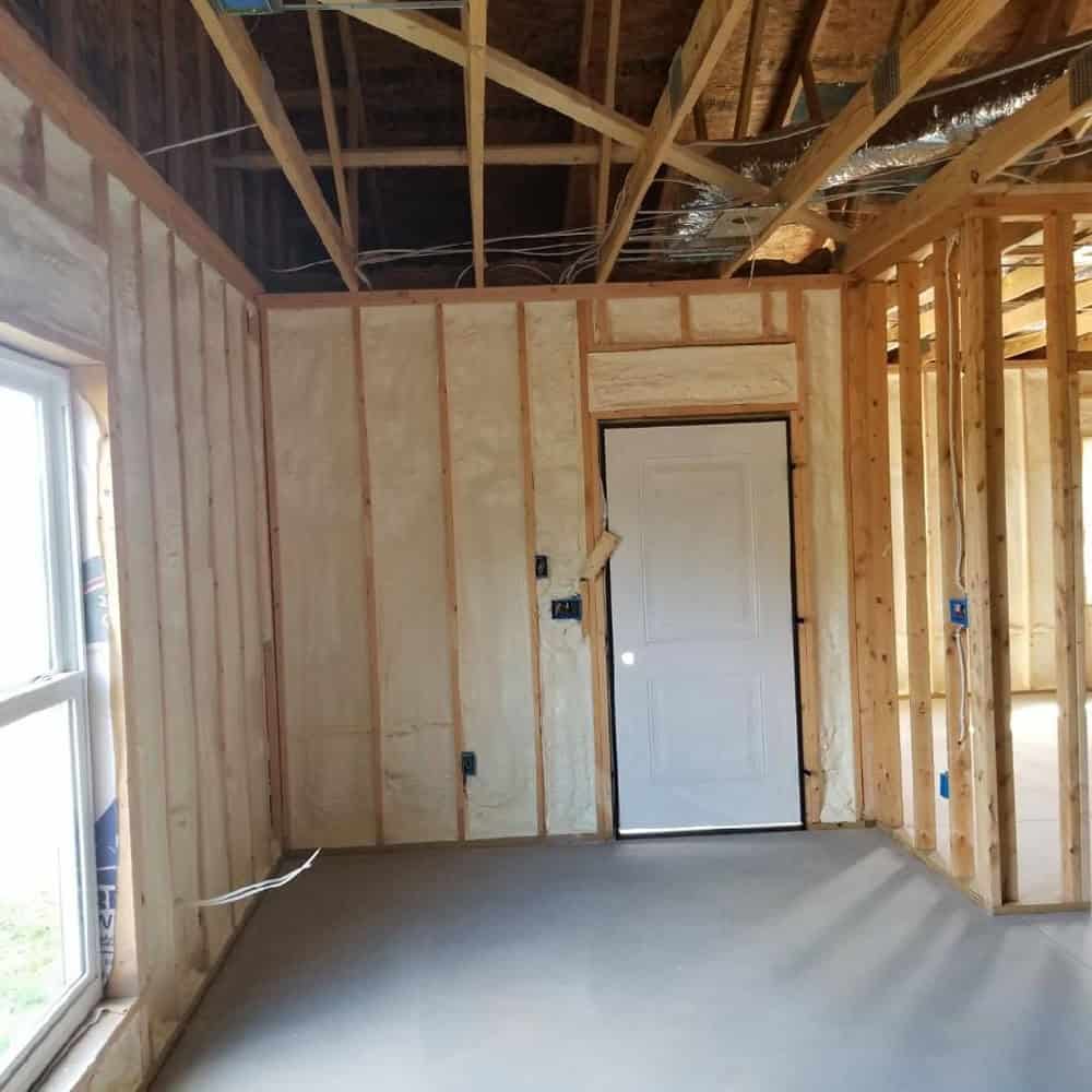 Room under construction with exposed wood framing, partially installed insulation, a closed white door, and a window letting in natural light. Ceiling beams and some wiring are visible. The floor appears unfinished.
