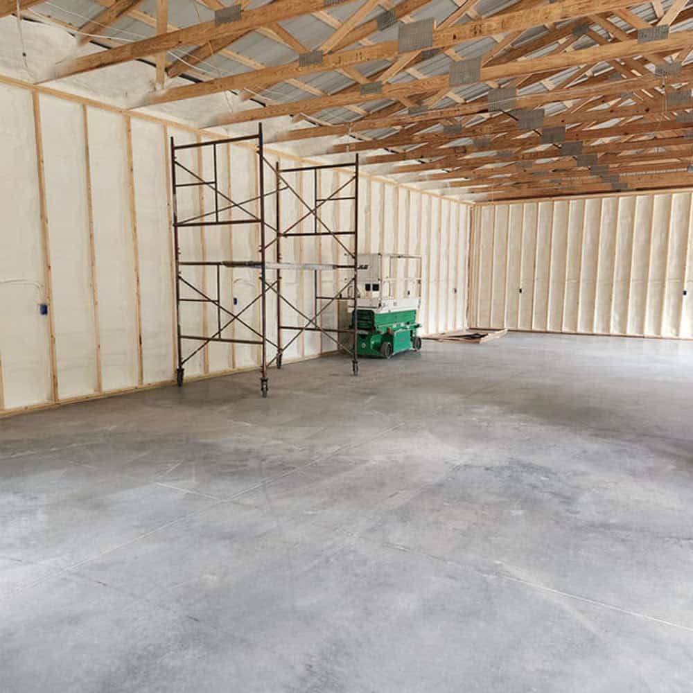 Interior of a large unfinished building with spray foam insulation on the walls, exposed wooden ceiling beams, concrete floor, a metal scaffold, and a green lift in the center.