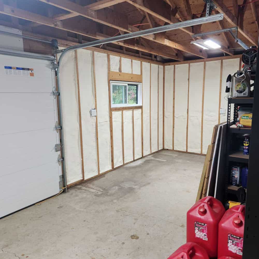 A garage with exposed wooden wall studs and spray foam insulation, a small window, concrete floor, and a partially open garage door. Red gas cans and shelves with various items are visible in the foreground.