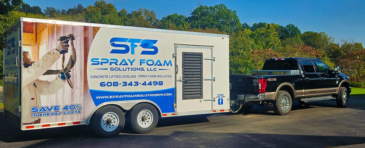 A black pickup truck tows a trailer with "STS Spray Foam Solutions" branding, contact info, and an image of a worker spraying insulation. The trailer advertises saving 40% in energy costs. Trees and blue sky are in the background. A black pickup truck tows a trailer with "STS Spray Foam Solutions" branding, contact info, and an image of a worker spraying insulation. The trailer advertises saving 40% in energy costs. Trees and blue sky are in the background.
